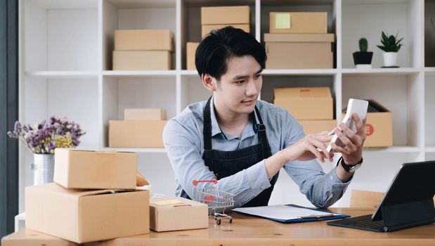 A portrait of Asian man, e-commerce employee sitting in the office full of packages on the table using a calculator, for SME business, e-commerce, technology and delivery business..