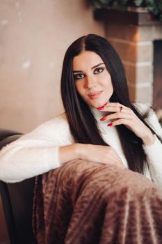 Portrait of stunning attractive young woman with long dark hair and bright evening make up in white sweater sitting on the floor with christmas gifts. She is holding white toy dwarf. Looking at camera.