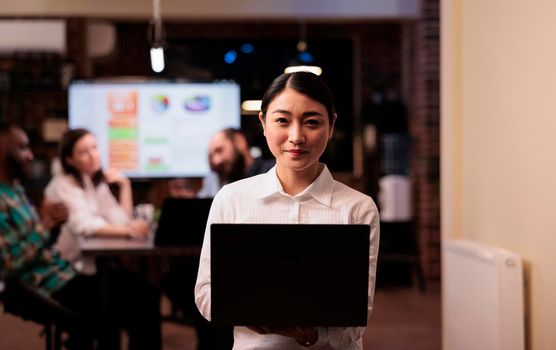 Portrait of smiling asian office worker holding laptop in business office during late night meeting working overtime. Startup employee posing confident in busy workspace with mixed team discussing sales charts.
