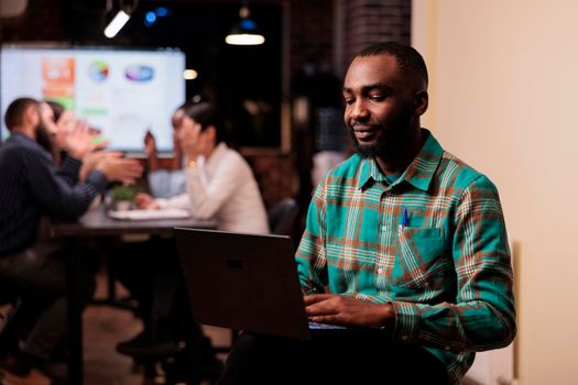 Portrait of smiling african american startup employee looking at laptop screen during late night meeting. Man working on portable computer posing confident with team discussing sales charts.