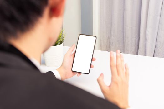 Work from home, Back view of Asian young businessman smile wearing suit video conference call or facetime by smart digital mobile phone blank screen raise hand to say hello teammates on desk