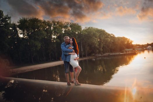 Beautiful full length portrait of young couple hugging at sunset by the river in park. Husband kissing his pregnant wife in the sunlight. Still surface of water reflects the sun.
