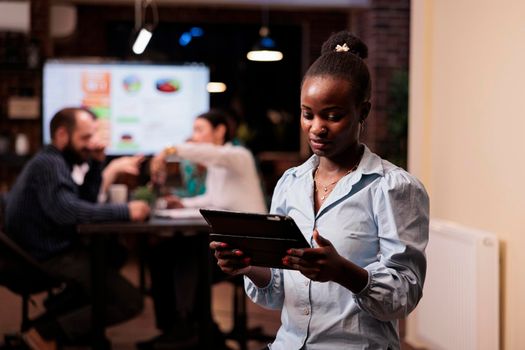 Focused african american woman holding tablet and looking at screen working late hours during business strategy meeting with mixed team. Startup employee posing confident in group project with coworkers.