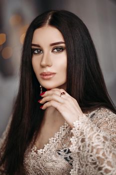 Portrait of stunning beauty with long dark hair and bright evening make up in lace sparkling dress looking at camera calmly and confidently. Bokeh.