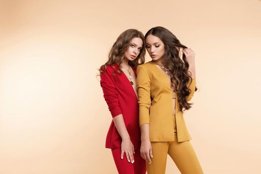 Full length side view of two elegant brunette ladies in stylish bright red and yellow suits and high heels giving high five to each other, smiling at camera. They are cooperating, working together or having an idea. Agreement concept.