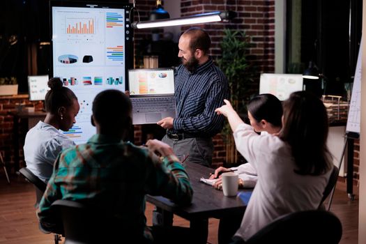 Office worker holding laptop with business presentation looking at screen while coworker is pointing at charts in late night meeting. Caucasian man standing in office talking with mixed team.