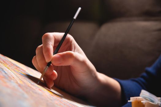 Close-up of a young female artist's hand with a thin brush painting a picture on canvas in a dark room. Shallow depth of field. high contrast.