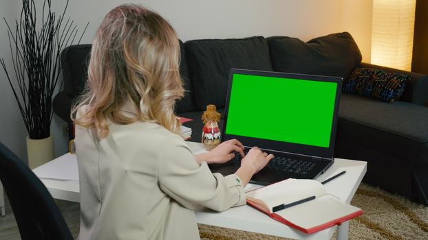 Blonde business woman sitting with her back in the gray jacket typing on the computer very focused. Green screen with color display. Working in office interior on laptop. Looking and sitting at table.