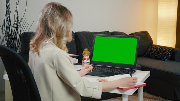 Back View of business Woman at Laptop with Green Screen for Copy Space. Chromakey Mockup. Home office work. Makes notes from laptop sitting on table. Blonde lady