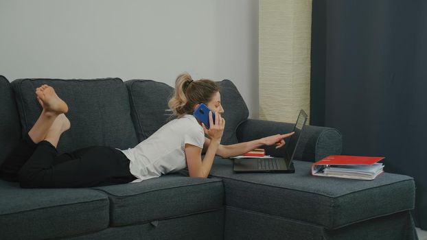 Young Caucasian woman sitting on couch in living room and discussing financial document with colleagues via phone call, points to the laptop screen. Concept project planning. Enthusiastic discussion.