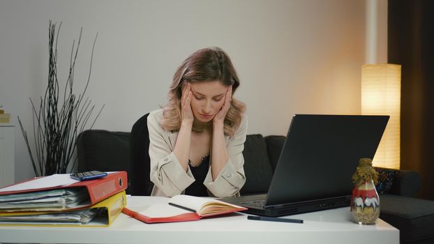 Tired woman, overwhelmed by laptop work, has severe headache. She takes a break while massaging her temples, is tired and feels unwell but tries to concentrate. Overworked woman with migraine.