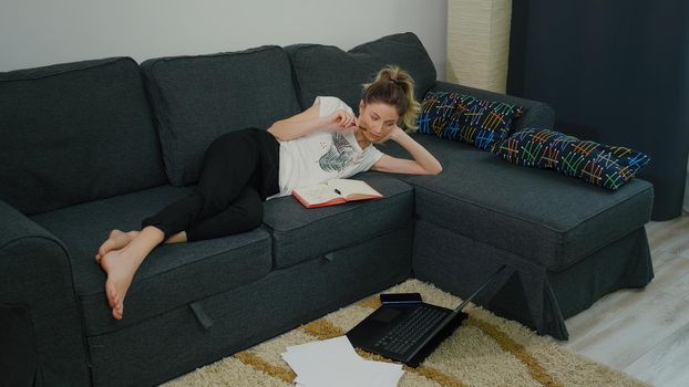 Student woman lying on the couch in the living room studying at home. Laptop is on the floor, necessary folders. Modern apartment large view. Boring study from home during pandemic period.