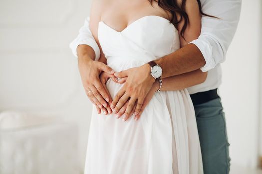Crop of tender husband holding his hands over his pregnant wife s hands over her belly. She is wearing beautiful sleeveless white wedding dress.