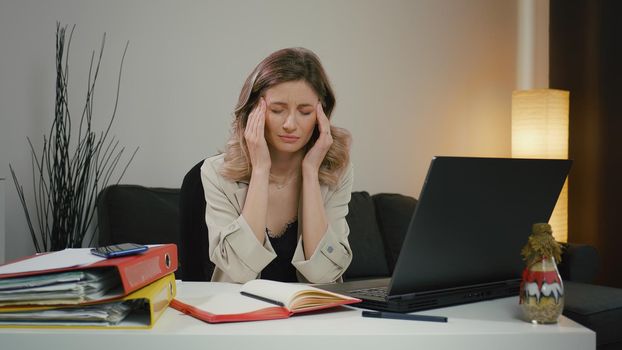 Tired woman, overwhelmed by laptop work, has severe headache. She takes a break while massaging her temples, is tired and feels unwell but tries to concentrate. Overworked woman with migraine.