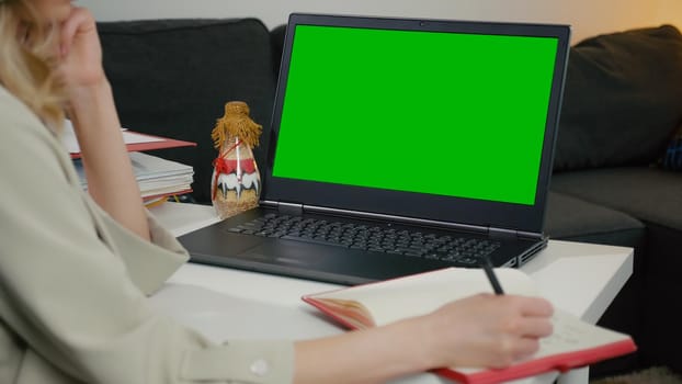 Close-up of Green Screen with Color Laptop Display. Businesswoman sitting at table takes notes in notebook from laptop, looking at screen and then at notebook. Working inside the home office.