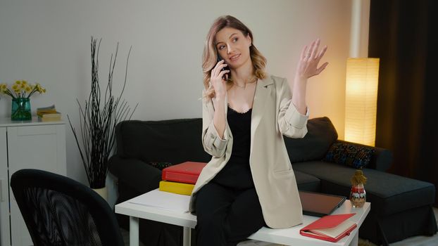 Happy Caucasian business woman explaining on the phone. She sits on the desk, cheerful, having a phone conversation. The concept of working from home and work misunderstandings. Gestures with hand.