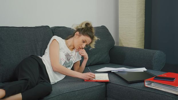 Pretty woman makes notes in notebook while lying on couch at home in a modern apartment. Do accounting in a notebook. Focused face. Write with pen in notebook. Concept working from home.