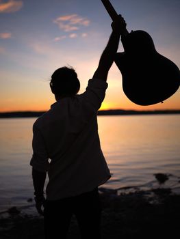 man raising his guitar on a lakeshore at nightfall, vertical silhouette close up view