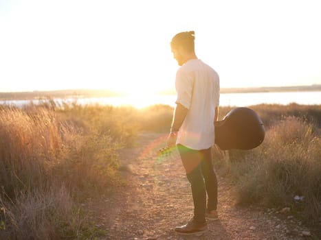 man standing pensively by the sunset in the middle of a field next to a lake, holding a guitar