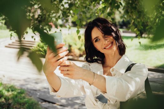 Beautiful Young Woman Making Selfie on Smartphone Outdoors Among Trees in Park at Summer Sunny Day