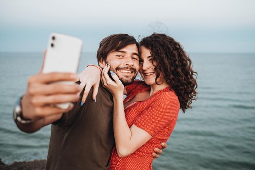 Happy Caucasian Couple Man and Curly Brunette Woman Hugging While Standing Against Sea and Making Selfie on Smartphone