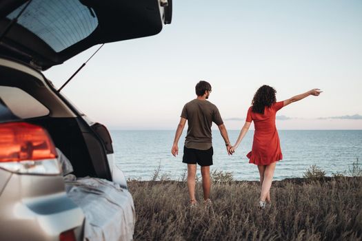 Young Couple Holding by Hands Walking on Background of the Sea, Man and Woman Enjoying Their Road Trip on Car