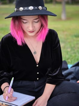 portrait of a young girl with pink hair and a black hat sitting with a notebook on her legs, blurred background