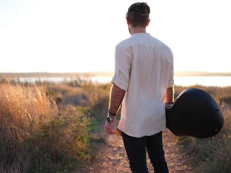 Musician contemplating the sunset with his guitar in his hand, standing on a path in front of a lake