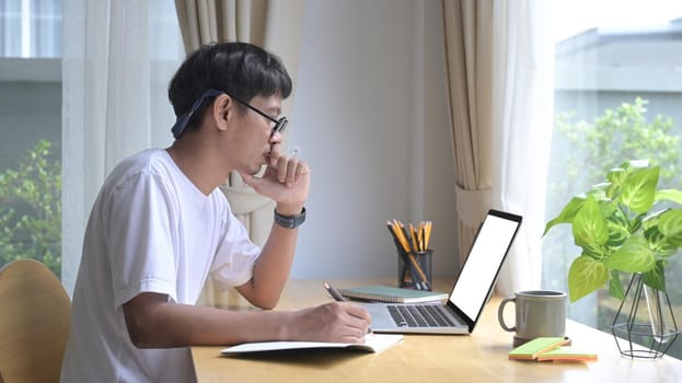 Focused asian man using laptop, reading email, working online at home office.