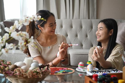 Positive mother and daughter painting Easter eggs together in living room.