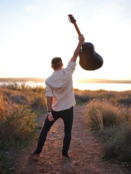 guitarist posing holding his guitar high in the air in front of a sunset by a lake, an expression of freedom in nature, vertical picture