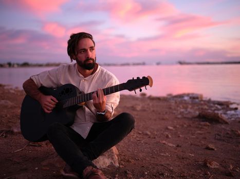 man sitting on a stone playing guitar on the shore of the pink lake at nightfall, horizontal portrait