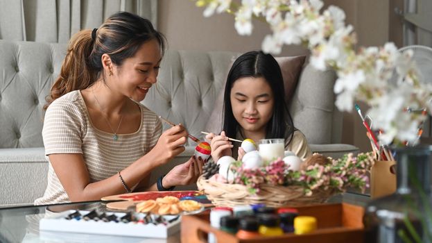 Smiling Asian mother and daughter painting Easter eggs together at home, family preparing for Easter.