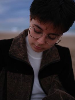 transgender woman with glasses and nose piercing resting looking at the ground, cut-out view with blurred beach in the background