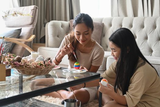 Happy asian mother and her daughter sitting in bright living room and painting Easter eggs together.