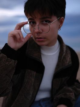 young trans woman taking off her glasses while looking intently at the camera, blurry sky in the background