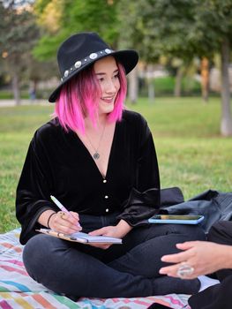 smiling girl with pink hair and hat sitting in the park writing in her notebook, horizontal portrait on blurred background