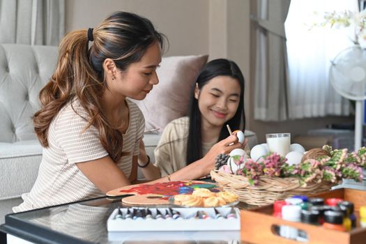 Beautiful asian mother helping her daughter painting easter eggs, family preparing for Easter.