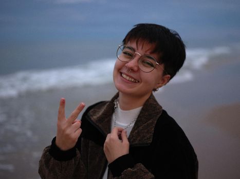 smiling transgender girl standing on the beach making victory sign with her hand, horizontal close up