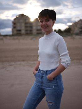 non-binary person posing smiling on the beach with hands in pockets, sunset in background