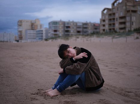 full view of a young transgender woman resting on the beach embracing herself, horizontal portrait