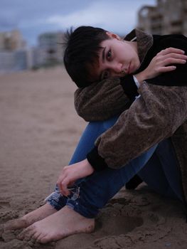 transgender woman sitting on the sand on the beach holding her glasses and with her knees bent