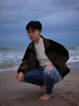 woman squatting barefoot posing on the seashore wearing long trousers and jacket, landscape vertical background