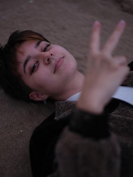 non-binary woman lying with her head on the sand, making a v-sign with her hand, vertical close-up