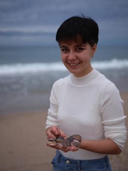 transgender looking at the camera with a white sweater and seashells in her hands, vertical portrait with the sea in the background.