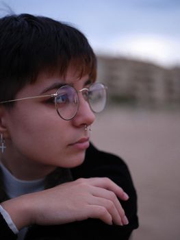 trans woman with glasses, piercings and short hair looking to the side, blurred background