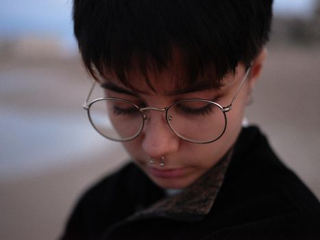 close-up view of the face of a young transgender girl with glasses and septum looking down, background out of focus