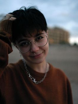 standing non-binary woman looking at the camera with one arm on her head, wearing glasses, a necklace and a brown jumper