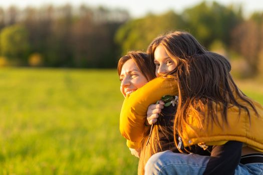 Playful mother giving daughter piggy back ride at green field. Both laughing and look happy. Spring in forest background. Closeup.