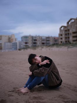 non binary young woman alone on the beach sitting on the sand looking at the camera, vertical portrait